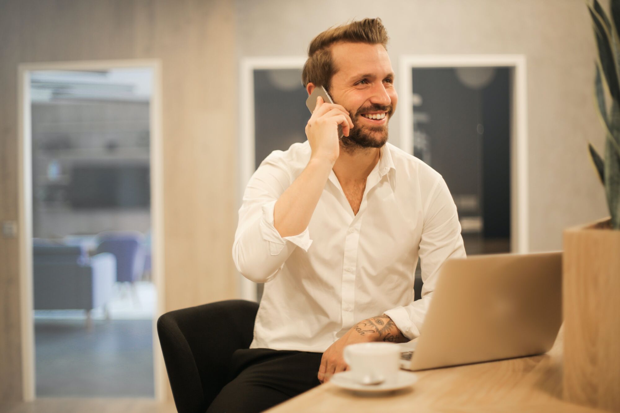 Homme d'affaires souriant au téléphone dans un bureau moderne avec un ordinateur portable et une tasse de café.