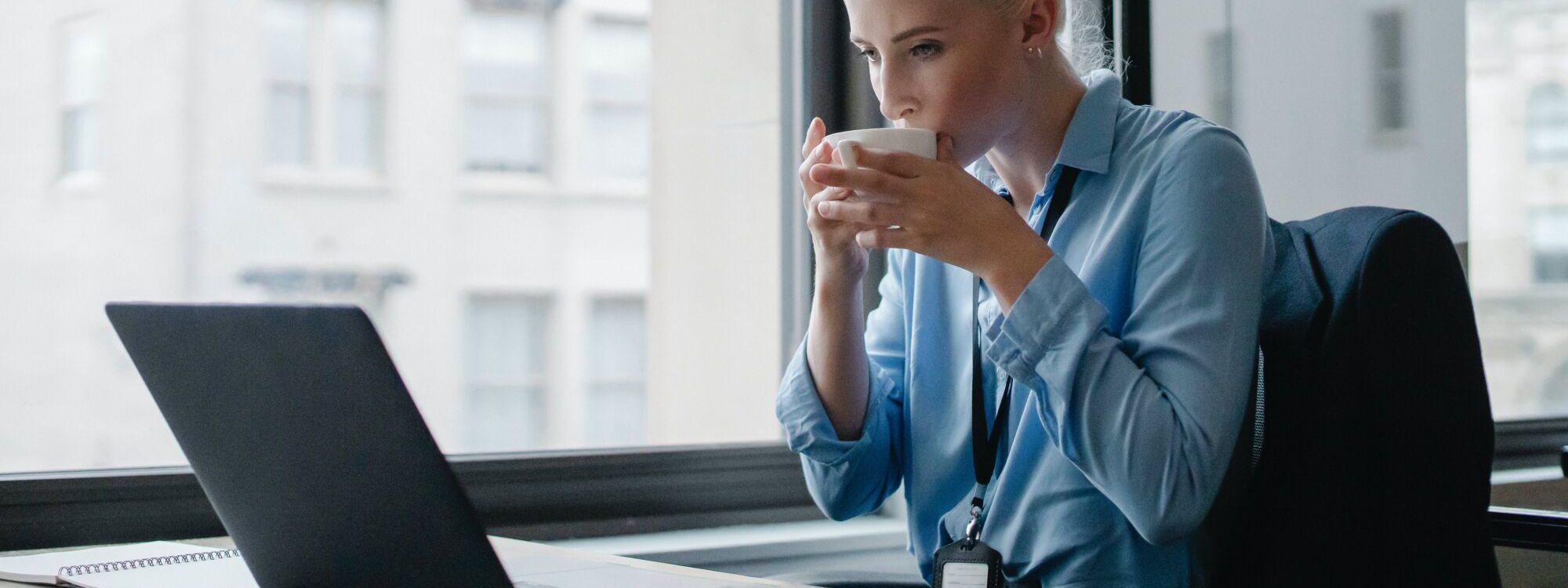 Femme d'affaires buvant une tasse de café tout en travaillant sur un ordinateur portable devant une fenêtre.