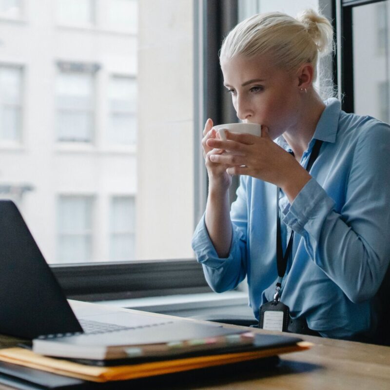 Femme d'affaires buvant une tasse de café tout en travaillant sur un ordinateur portable devant une fenêtre.