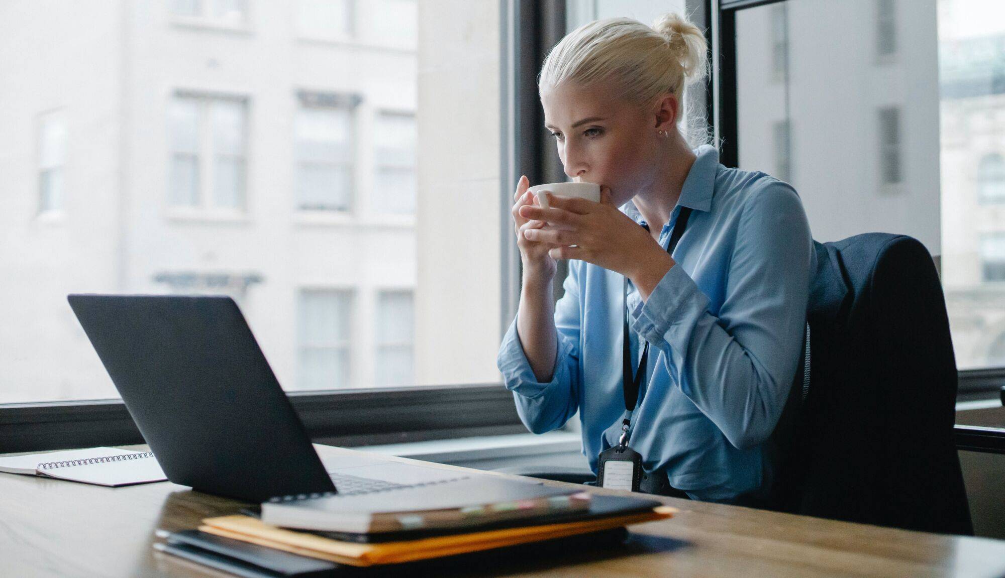 Femme d'affaires buvant une tasse de café tout en travaillant sur un ordinateur portable devant une fenêtre.