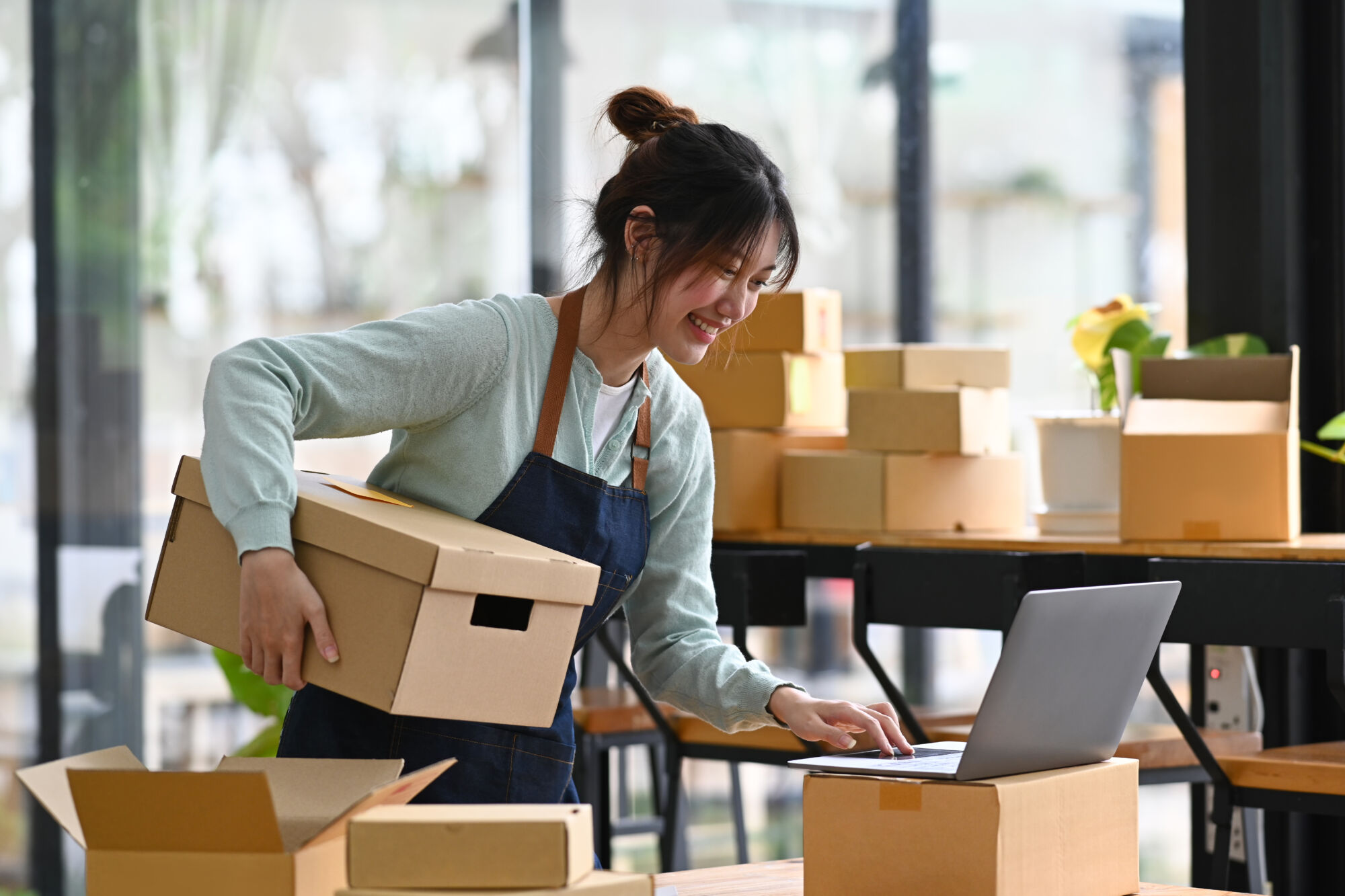 Femme souriante préparant des commandes tout en interagissant avec un ordinateur portable dans un espace de travail lumineux.