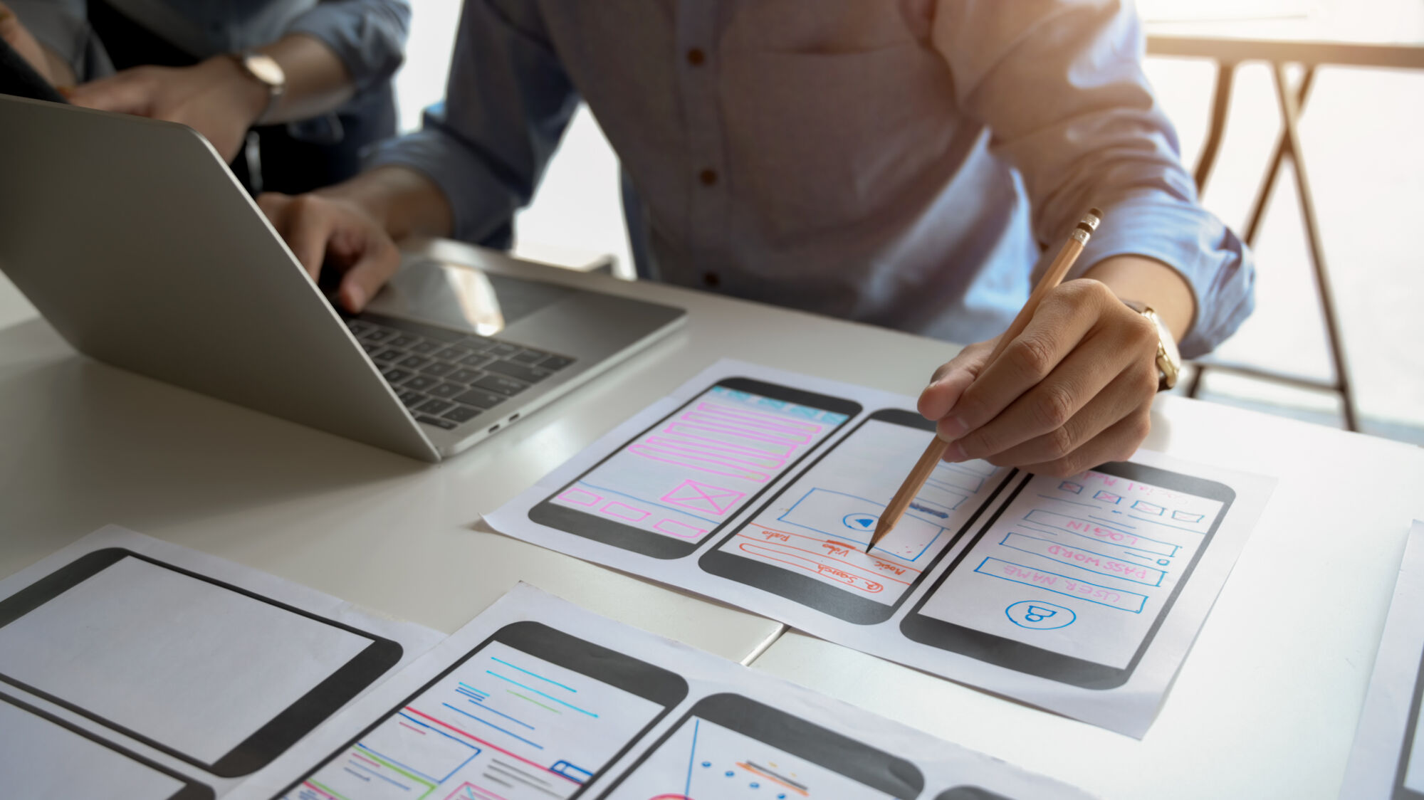 Personnes analysant des ébauches de conception d'interface mobile avec un ordinateur portable en arrière-plan.