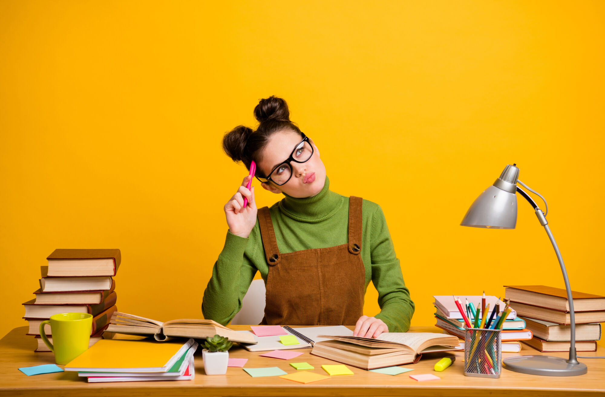 Étudiante réfléchissant entourée de livres et de notes avec un crayon rose à la main sur fond jaune.