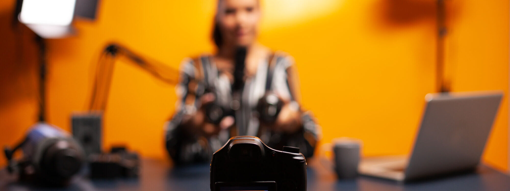 Femme réalisant une vidéo avec une caméra sur trépied dans un studio avec un fond orange et du matériel de tournage.