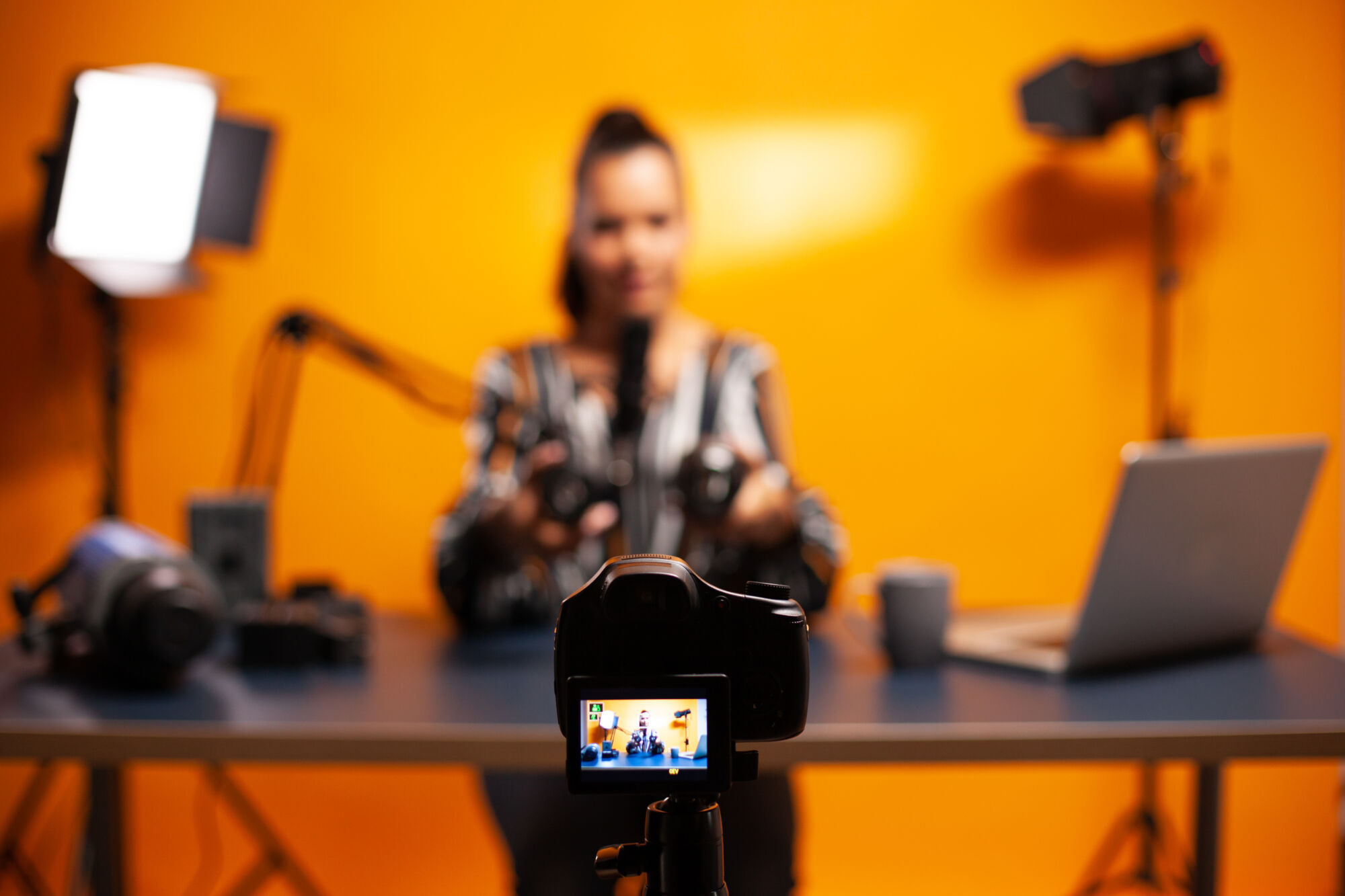 Femme réalisant une vidéo avec une caméra sur trépied dans un studio avec un fond orange et du matériel de tournage.