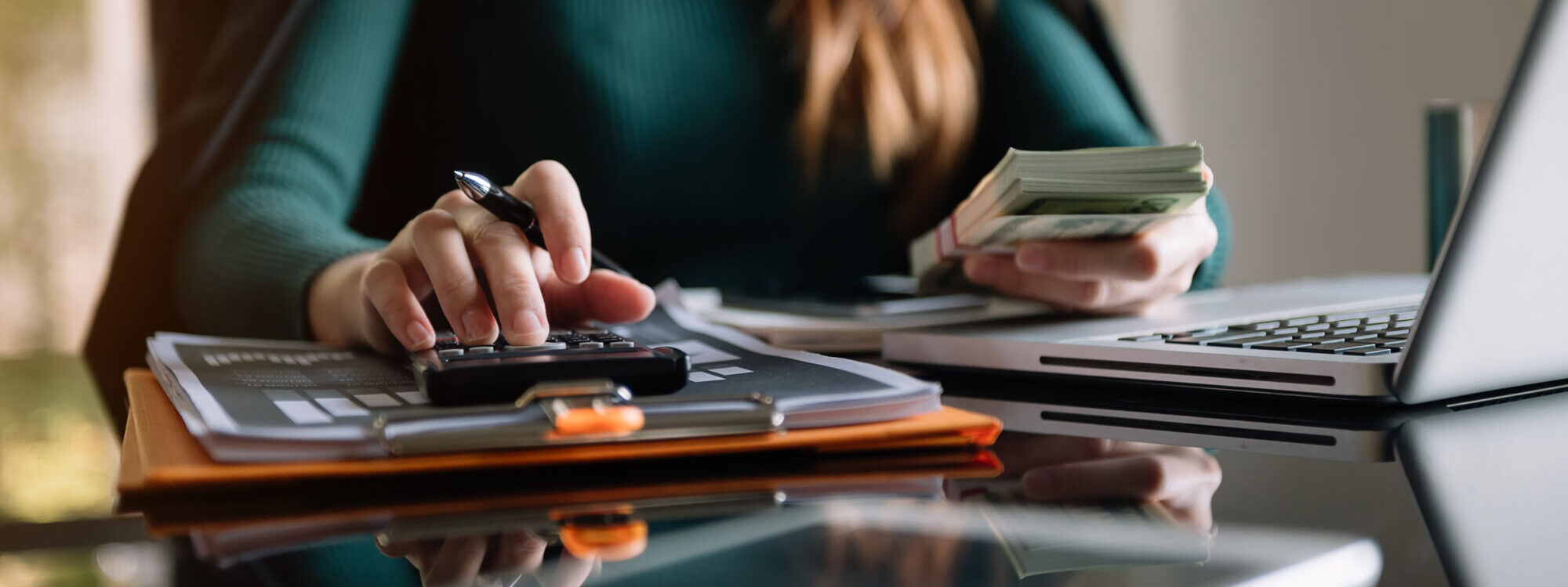 Femme calculant des finances avec un stylo, une pile de billets, une calculatrice et un ordinateur portable sur un bureau en verre.