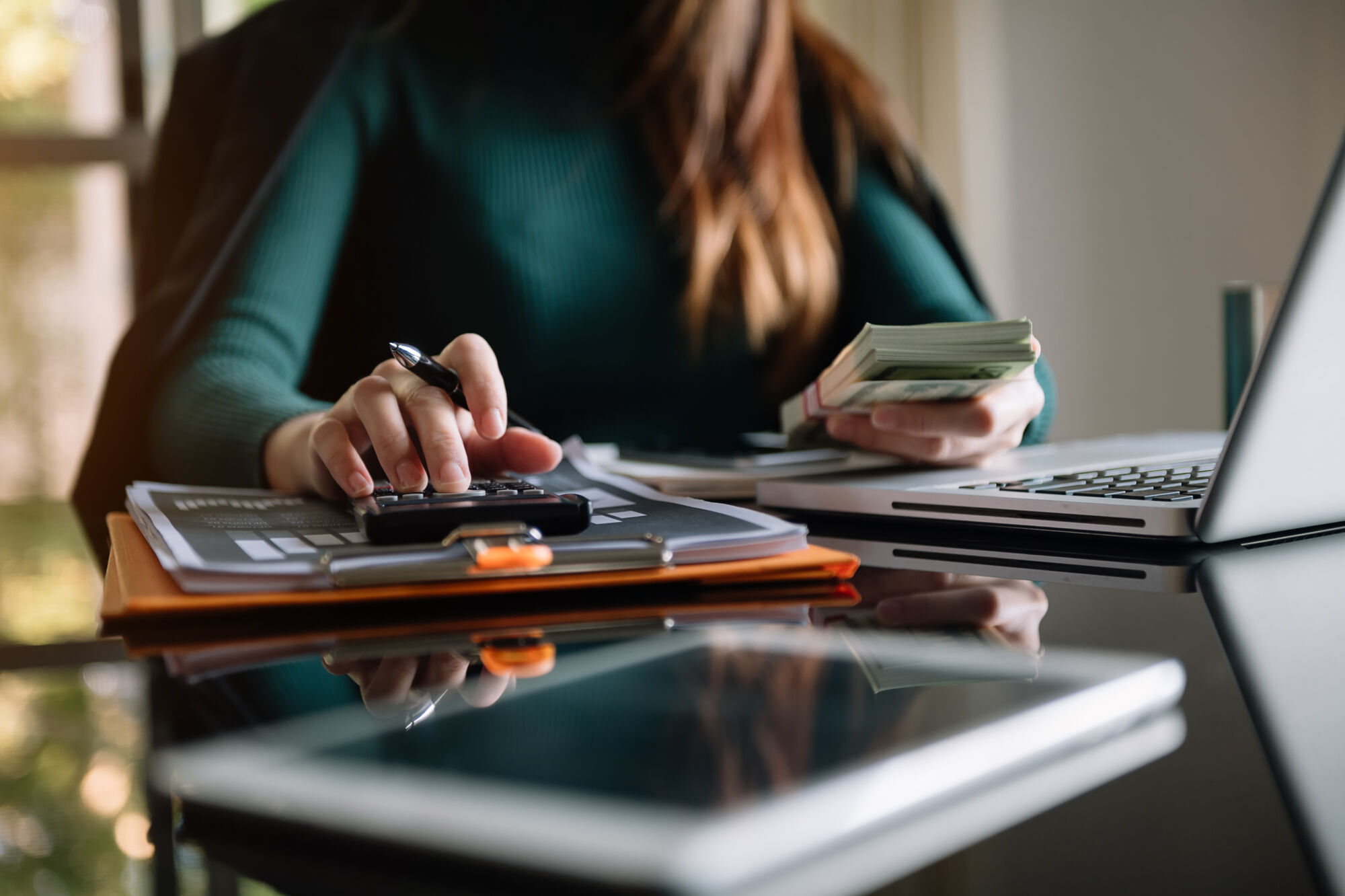 Femme calculant des finances avec un stylo, une pile de billets, une calculatrice et un ordinateur portable sur un bureau en verre.