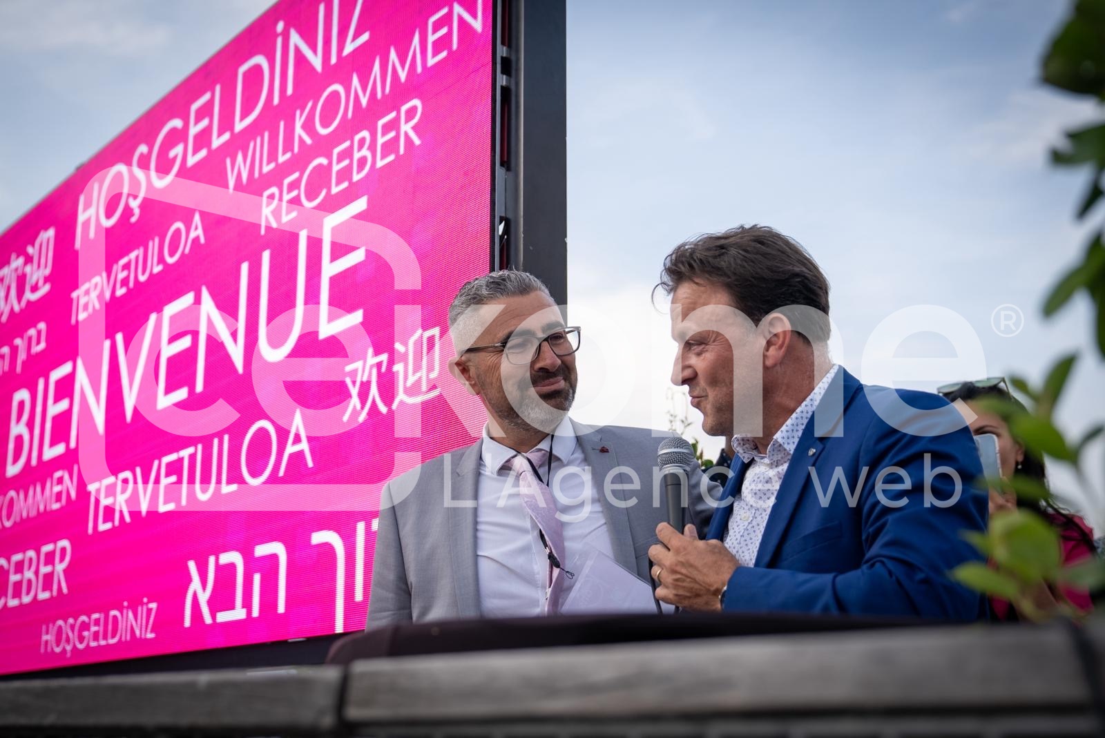 Deux hommes en conversation devant un écran d'affichage avec message de bienvenue multilingue lors d'un événement Cekome.