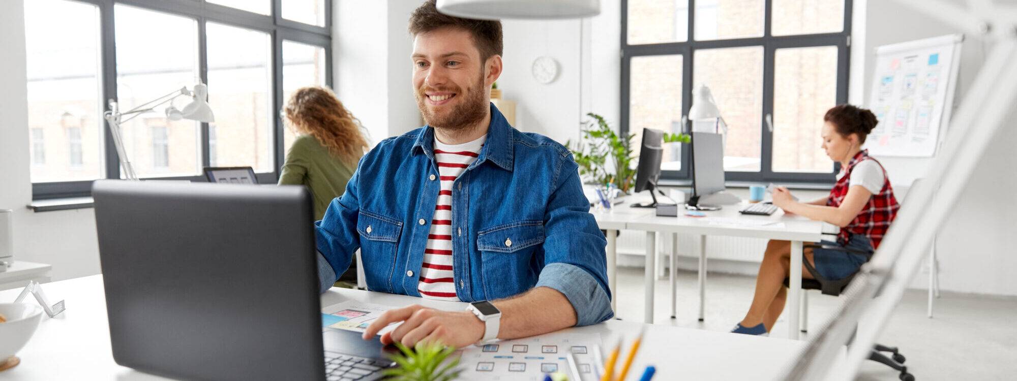 Homme souriant travaillant sur un ordinateur portable dans un espace de bureau moderne avec des collègues en arrière-plan.