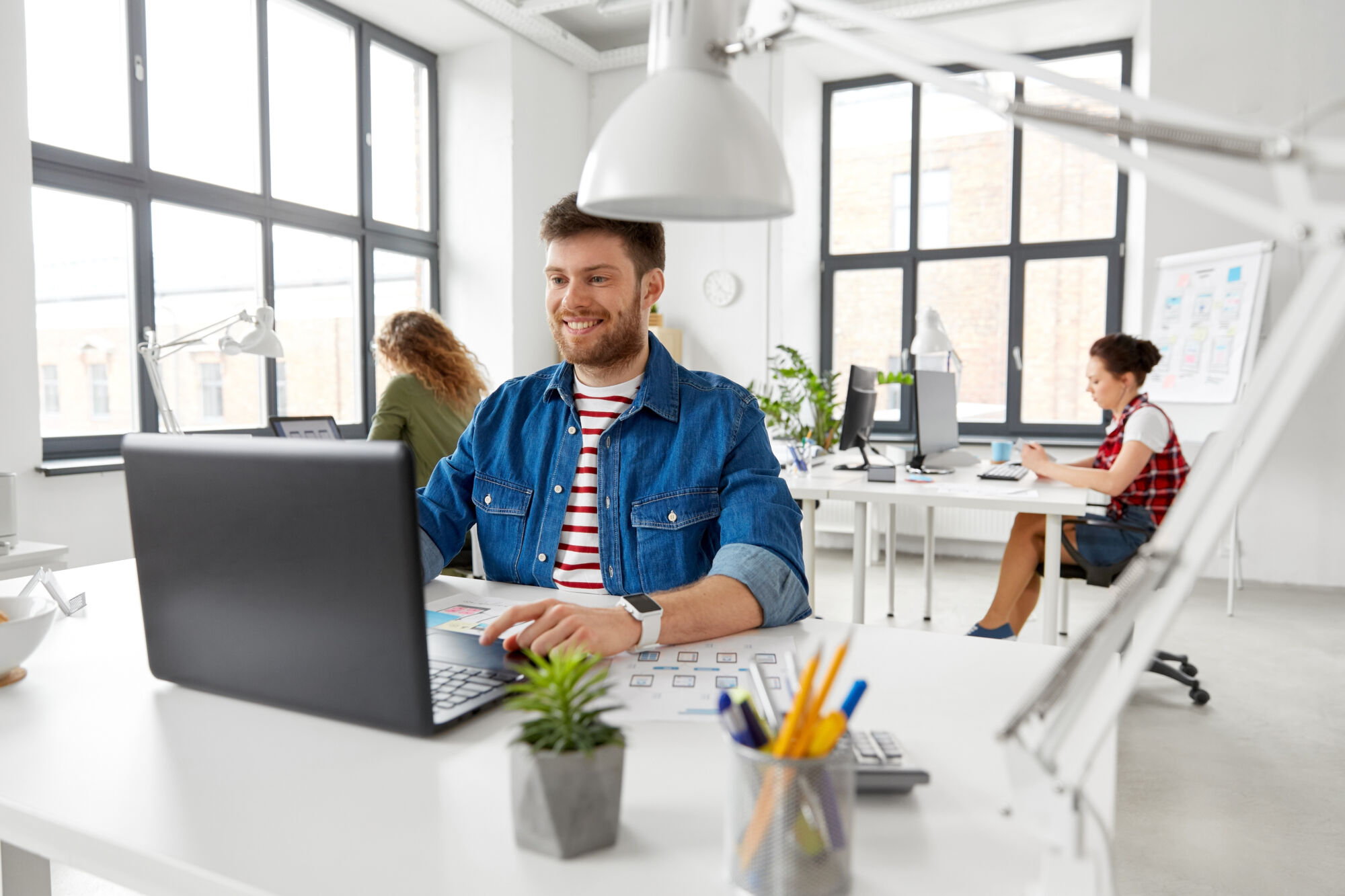 Homme souriant travaillant sur un ordinateur portable dans un espace de bureau moderne avec des collègues en arrière-plan.