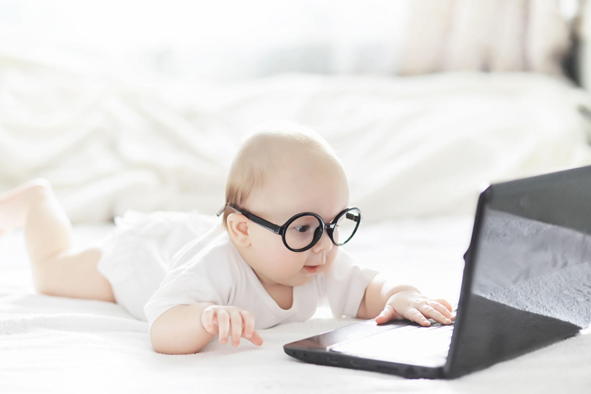 Bébé avec des lunettes regardant un ordinateur portable ouvert sur un lit, concept d'apprentissage précoce ou de technologie.