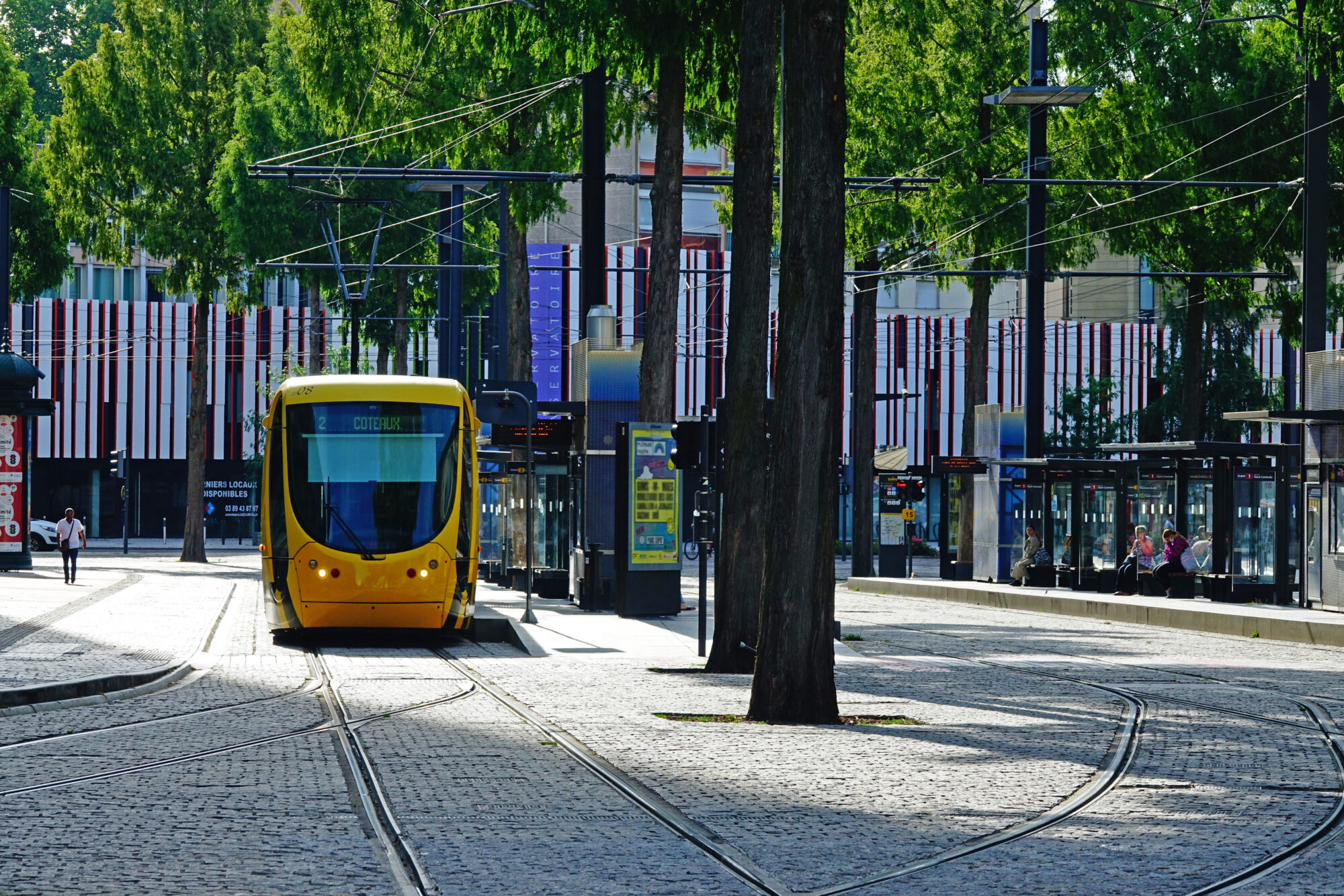 Tramway jaune dans une rue bordée d'arbres avec station de tramway moderne.