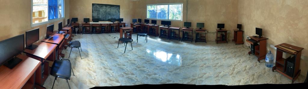 Salle de classe vide avec des bureaux et des ordinateurs, lumière naturelle venant de la fenêtre.