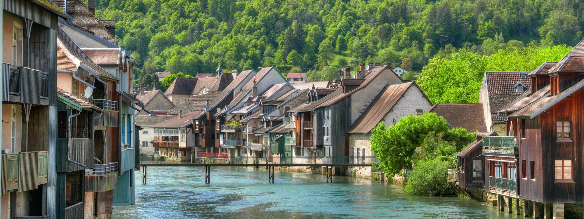 Maisons traditionnelles au bord de l'eau dans un village avec collines verdoyantes en arrière-plan.