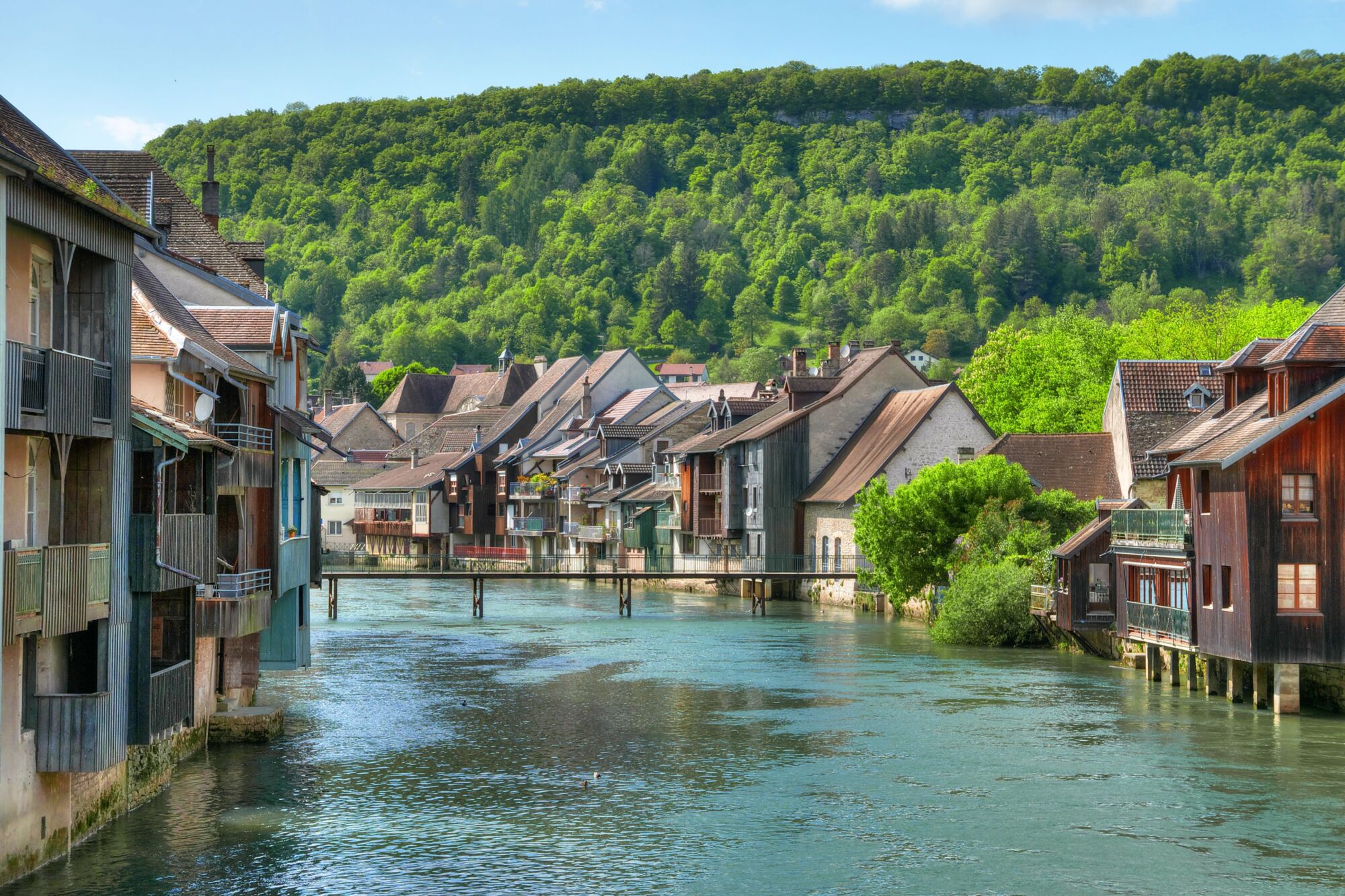 Maisons traditionnelles au bord de l'eau dans un village avec collines verdoyantes en arrière-plan.