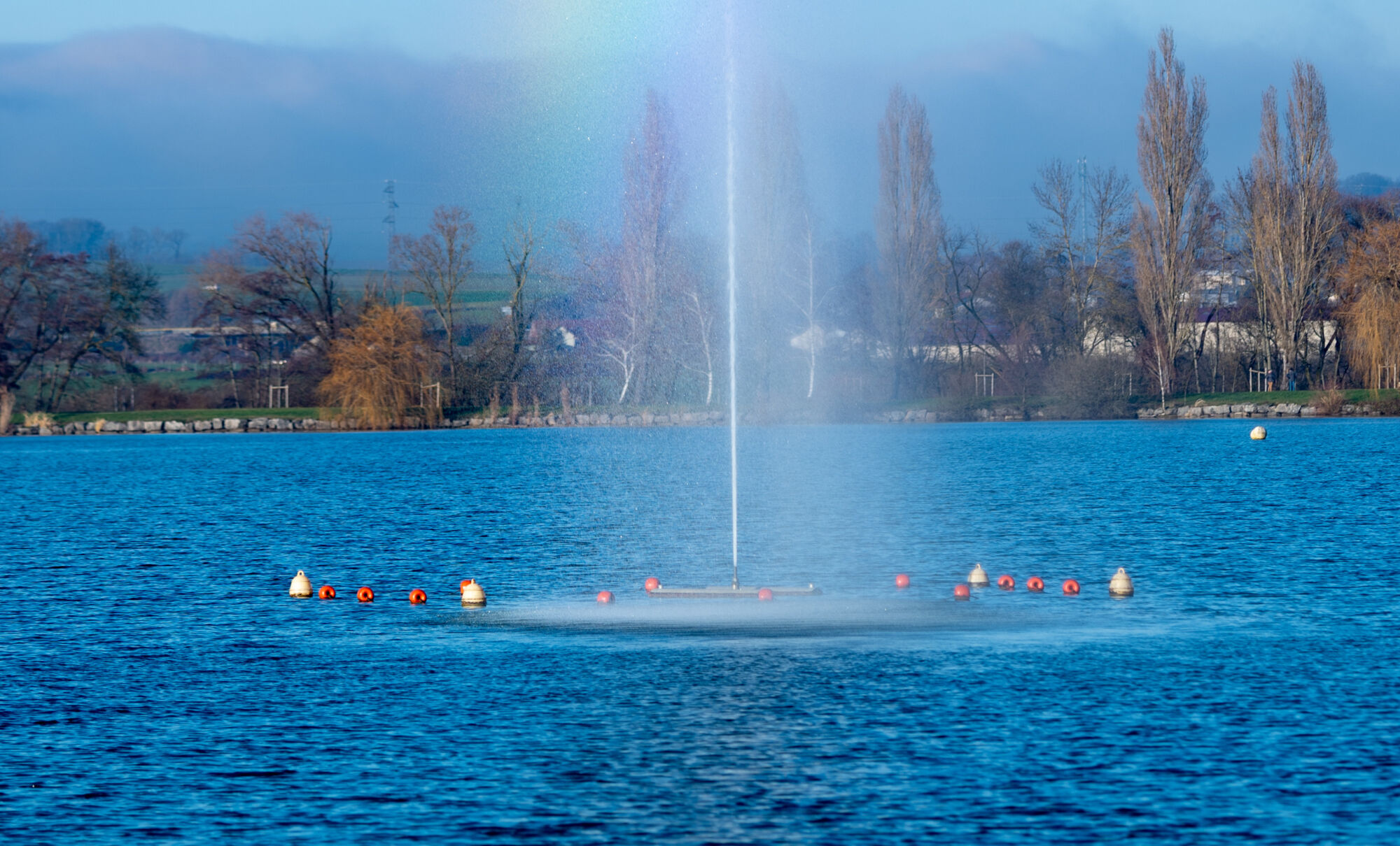 Jet d'eau dans un lac avec arbres et montagnes en arrière-plan sous un ciel bleu.