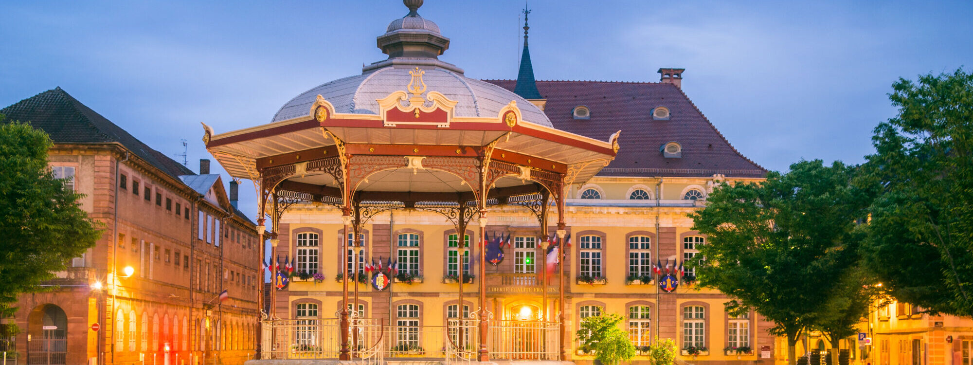 Kiosque à musique historique éclairé au crépuscule avec bâtiment municipal en arrière-plan.