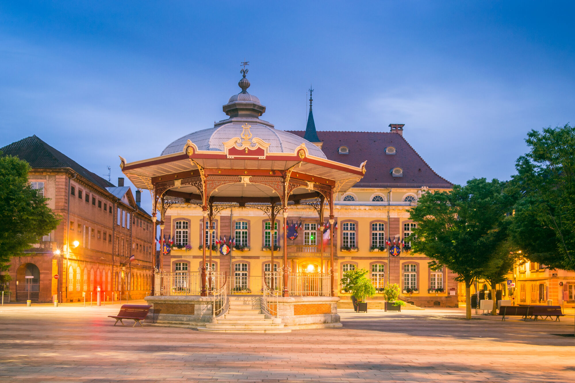 Kiosque à musique historique éclairé au crépuscule avec bâtiment municipal en arrière-plan.