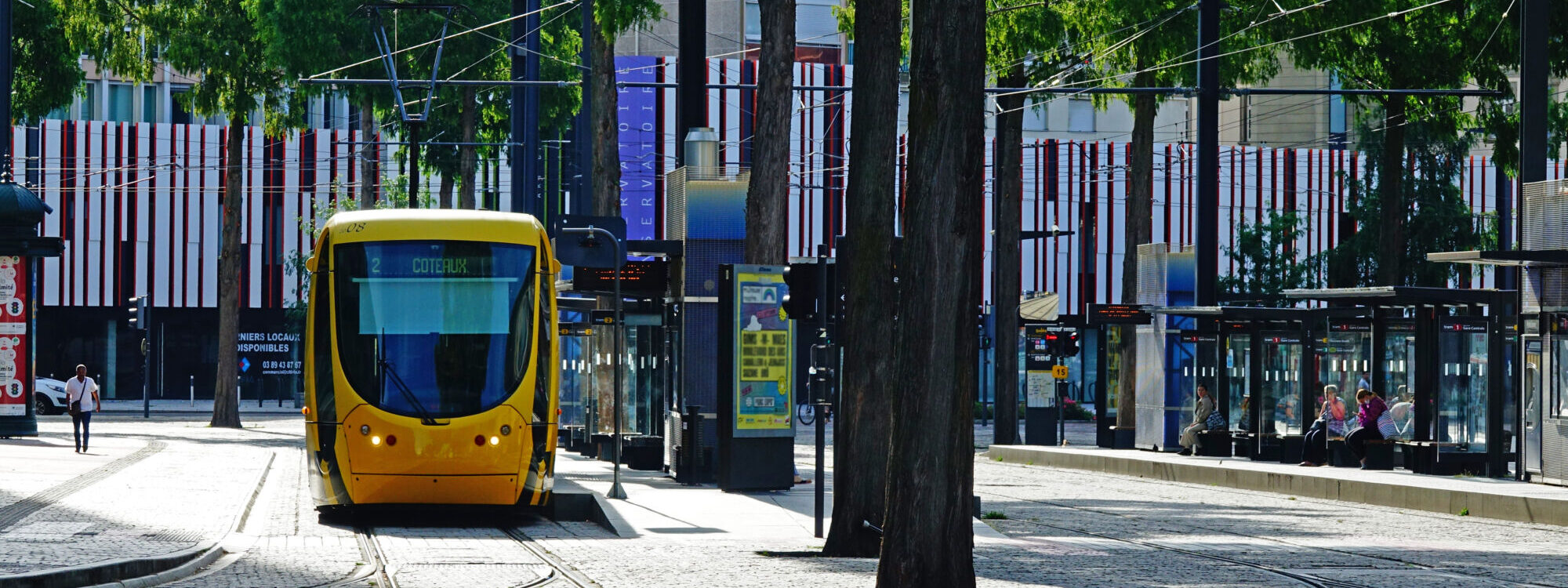 Tramway jaune dans une rue bordée d'arbres avec station de tramway moderne.