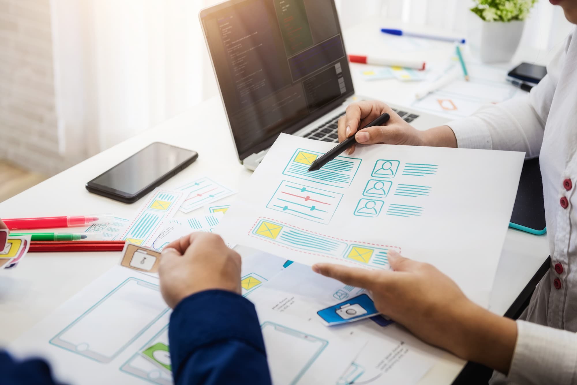 Deux professionnels analysant des maquettes de design d'interface utilisateur sur une table avec des papiers et un ordinateur portable.