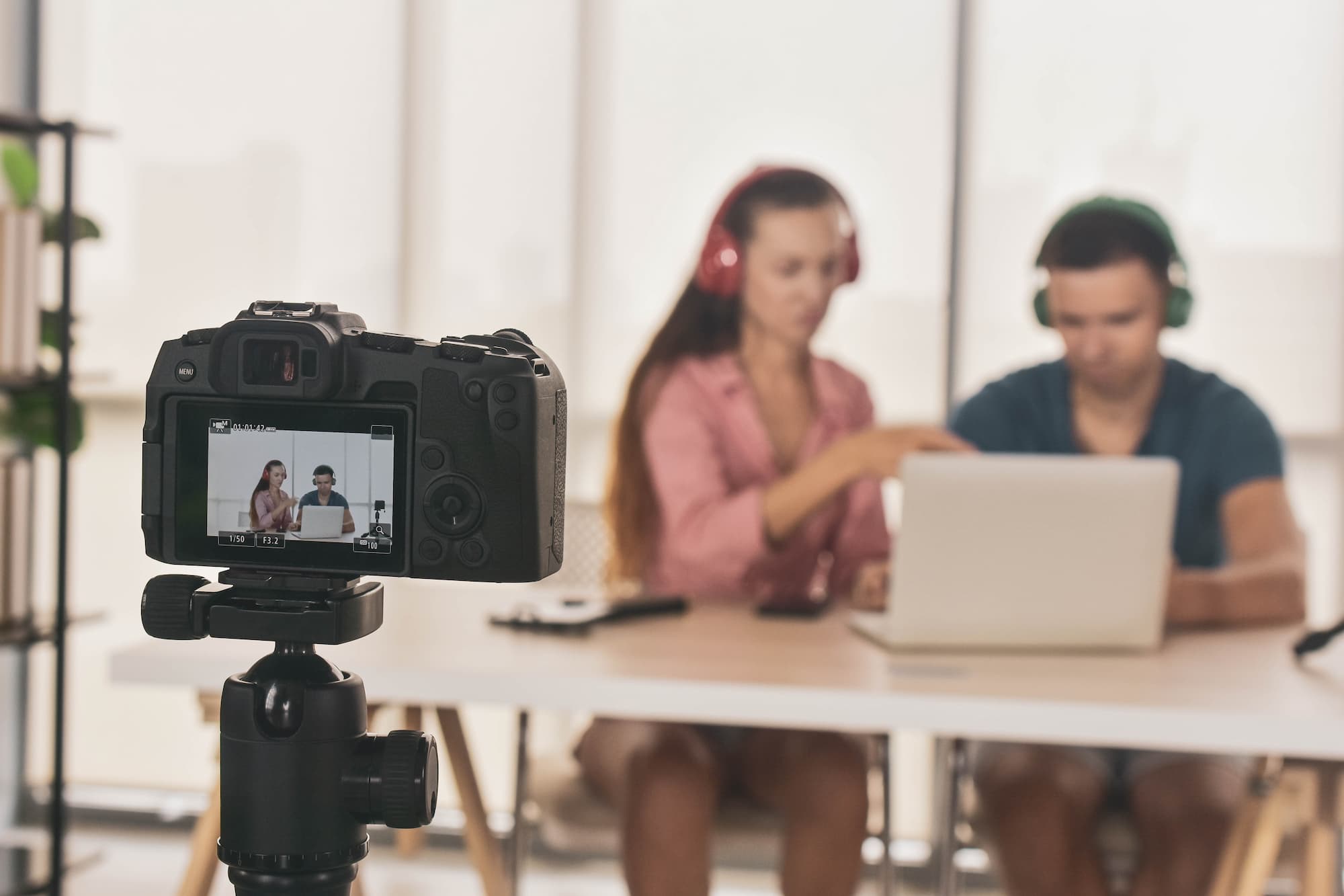 Deux personnes collaborant et filmées par une caméra sur un trépied, avec des écouteurs et un ordinateur portable.