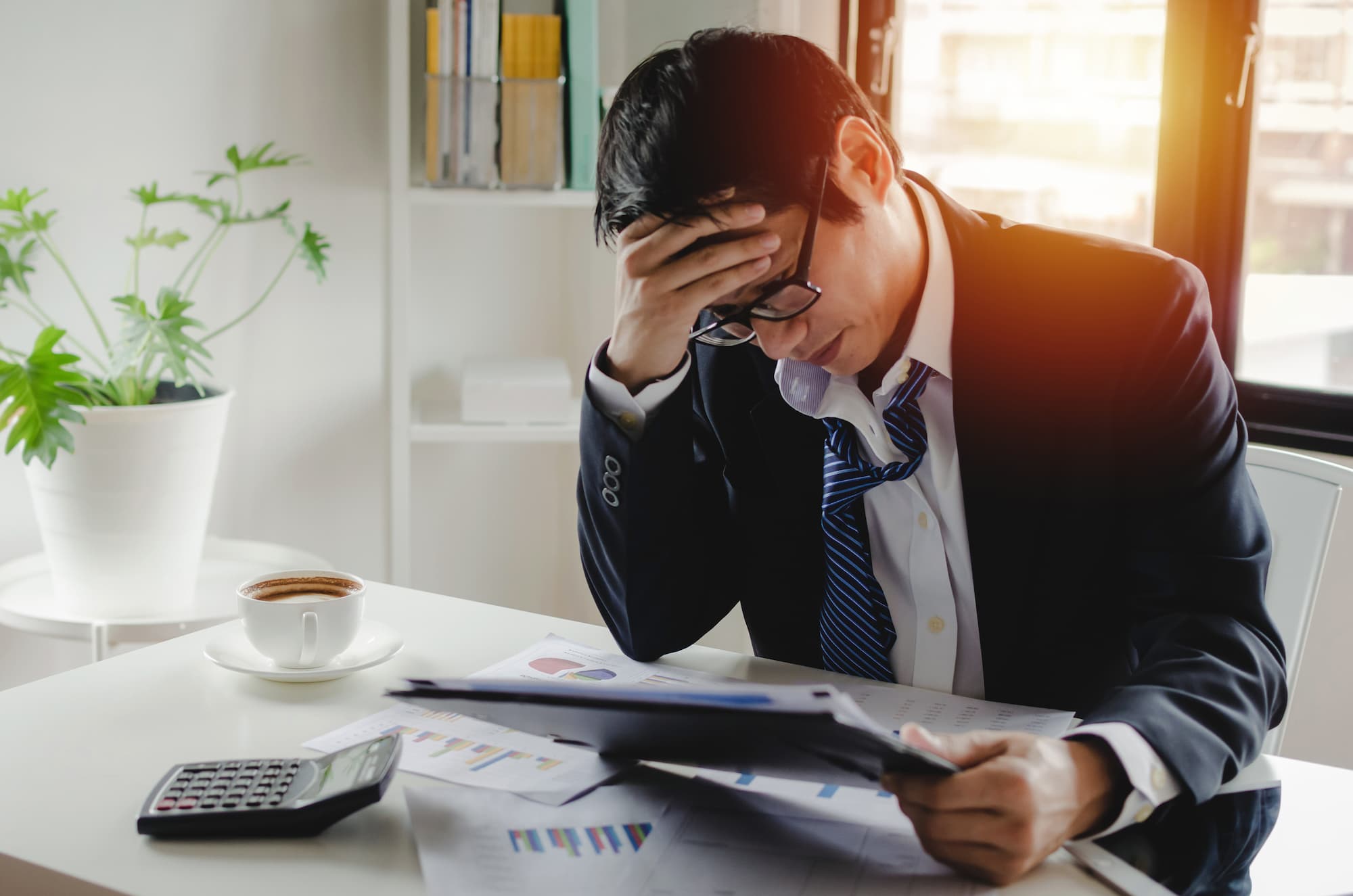 Homme d'affaires frustré tenant sa tête au-dessus d'un rapport financier dans un bureau lumineux avec une tasse de café et une calculatrice