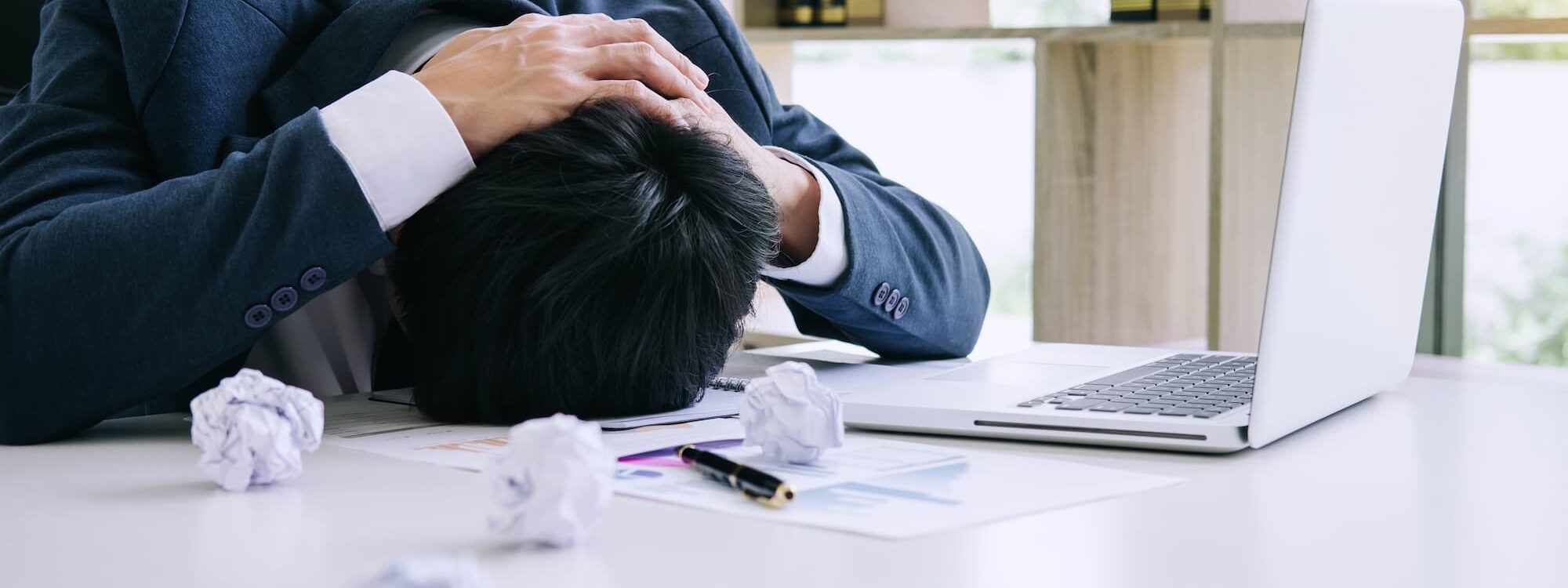 Homme d'affaires asiatique épuisé dormant sur son bureau encombré de papiers froissés, devant un ordinateur portable ouvert