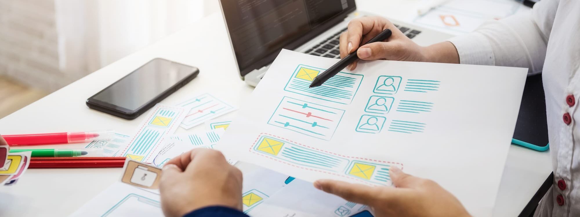 Deux professionnels analysant des maquettes de design d'interface utilisateur sur une table avec des papiers et un ordinateur portable.