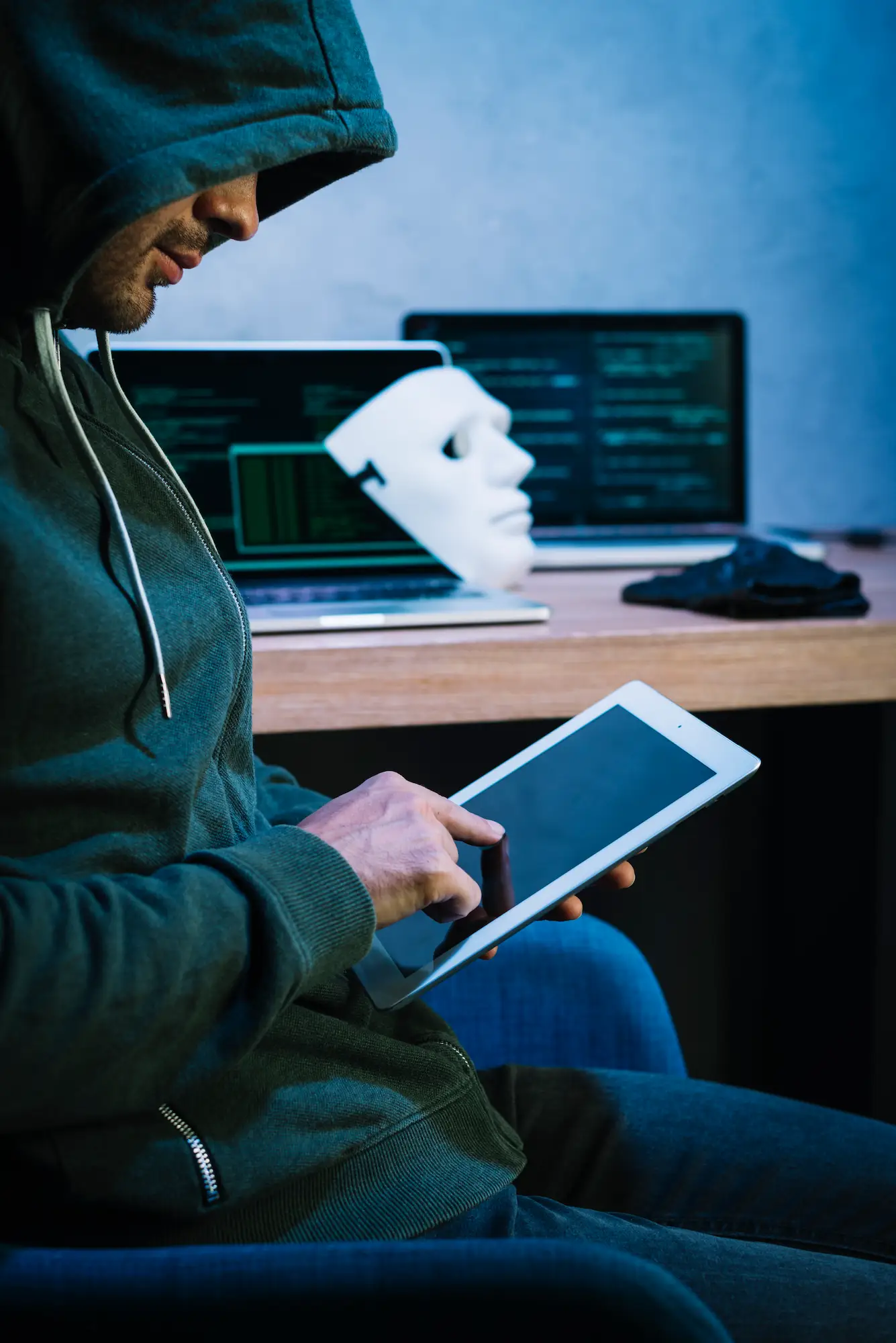 Personne portant un sweat à capuche utilisant une tablette, avec un masque blanc et des écrans affichant du code en arrière-plan